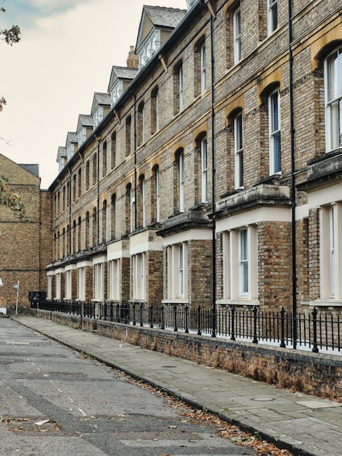 Photograph of a quiet residential street in Kentish Town featuring a row of terraced brick and painted houses with small front gardens and railings. The street is paved with cobblestones and lined wit
