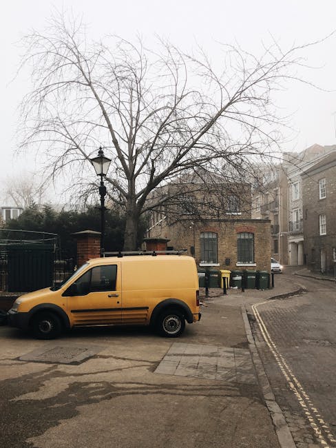 A yellow commercial van parked on a paved street in a residential area with brick buildings and a leafless tree nearby. The street features dual yellow lines along the curb, indicating parking restric