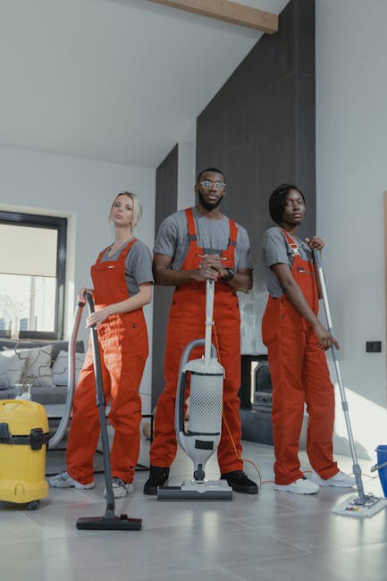 Three professional cleaners from Cleaners Kentish Town dressed in grey shirts and red overalls are standing in a modern living room, each holding a cleaning tool. The first cleaner on the left is holding a vacuum cleaner nozzle, while the center cleaner is holding a handheld vacuum with a cord, and the third cleaner on the right is holding a mop. The room features a polished concrete floor, a black accent wall, and a large window allowing natural light to illuminate the space. Visible cleaning equipment includes a yellow wet-dry vacuum and a blue dustpan with brush, indicating a comprehensive cleaning operation. The scene demonstrates thorough surface cleaning and hygiene maintenance typical of deep cleaning services offered by [COMPANY_NAME], emphasizing high standards of cleanliness in residential environments, aligning with the page about trusted deep cleaning services in Kentish Town.