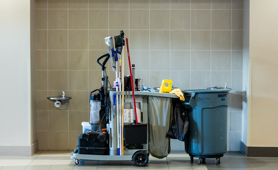 A cleaning trolley filled with various cleaning tools and supplies, positioned against a beige tiled wall in a well-lit room. The trolley holds floor mops, a vacuum cleaner, spray bottles, cloths, and other cleaning equipment. To the right of the trolley is a large blue waste bin with a black plastic bag lining, and to the left, a wall-mounted stainless steel sink. The surfaces, including the floor and tiles, appear clean and dry, reflecting a high standard of hygiene. The image illustrates professional domestic cleaning by Cleaners Kentish Town, emphasizing surface cleaning and sanitisation processes within a residential or commercial setting.